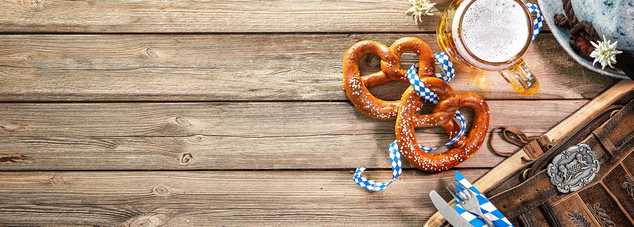 Bayerische Oktoberfest-Artikel: Brezeln, Bierkrug, Hut und Lederhosen auf rustikalem Holztisch.