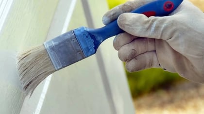 Una mano enguantada sostiene un pincel de mango azul, aplicando pintura blanca sobre una superficie blanca al aire libre.