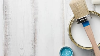 Lata de pintura blanca, pincel, pintura azul y rodillo sobre una superficie de madera blanca desgastada.