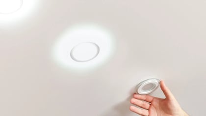 Mano instalando un downlight LED empotrable redondo blanco en el techo; dos otras luces encendidas son visibles.