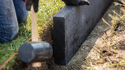 Ein grauer Gartenrandstein wird mit einem Gummihammer in den Boden eingesetzt, um einen sauberen Übergang zum Rasen zu schaffen.