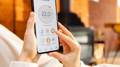 Manos sostienen un smartphone mostrando una app de hogar inteligente para controlar la temperatura (22.0°C) y otros dispositivos en la sala.