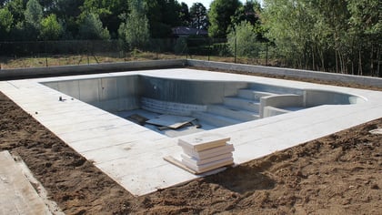 Ein im Bau befindlicher Außenpool mit hellen Pflastersteinen, Betonstufen und Innenwandpaneelen, umgeben von Erde.