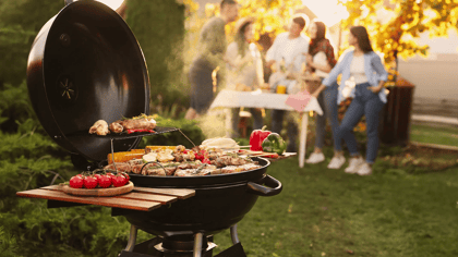 Schwarzer Kugelgrill mit Fleisch und Gemüse, im Freien bei einer lebhaften Gartenparty in Gebrauch.