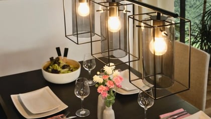 Lámpara colgante de metal negro con tres pantallas de cristal, iluminando una mesa de comedor.