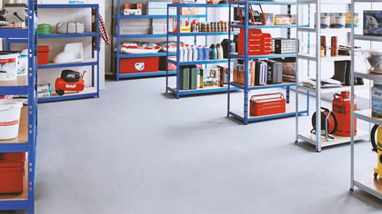 Toma de un almacén con estanterías azules y grises llenas de herramientas y materiales, incluyendo cubos de pintura, cajas de herramientas y otros artículos.