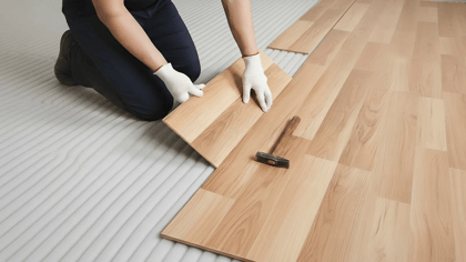 Ein Mann verlegt einen hellen Holzlaminatboden mit Hammer, weiße Handschuhe und grauer Unterlage für eine optimale Schalldämmung.