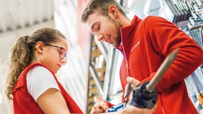 Empleados de tienda con uniformes rojos examinan herramientas manuales, con herramientas eléctricas naranjas y negras expuestas en estantes.