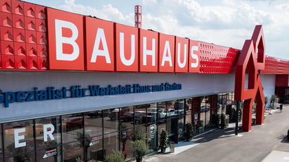 Großes rotes BAUHAUS Geschäft mit weißem Logo und blauem Text, mit Parkplatz, Autos und Fußgängern.