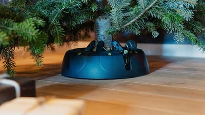 Schwarzer Weihnachtsbaumständer mit Fußpedal hält einen echten Baum auf Holzboden mit Geschenken.