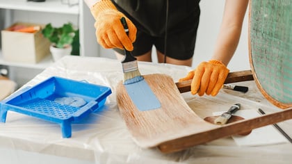 Person mit orangefarbenen Handschuhen streicht einen hölzernen Stuhlsitz hellblau mit einem Pinsel und einer blauen Farbschale.