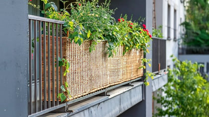Wunderschöner Balkonkasten aus Weide mit üppigen Pflanzen und bunten Blumen auf einem grauen Balkon. Natürliche Balkongestaltung.