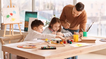Ein Mann und zwei Kinder sitzen an einem Tisch und malen mit bunten Farben und Pinseln. Das Bild zeigt eine kreative und künstlerische Szene.