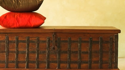 Un baúl decorativo de madera oscura con correas metálicas, con un cojín rojo y uno marrón encima, frente a una pared amarilla clara.