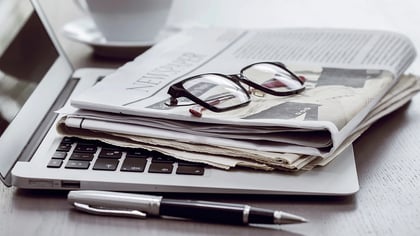 Ein Laptop mit einer Zeitung, schwarzer Brille und einem Stift auf einem Holzschreibtisch. Eine weiße Kaffeetasse ist im Hintergrund.