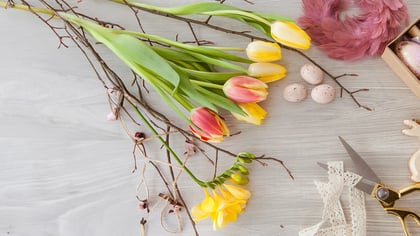 Frühlingshafte Osterdekoration mit Tulpen, Ostereiern, Federn und dekorativen Elementen auf hellem Holzgrund.