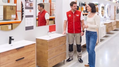 Dos personas discuten muebles de baño de madera con lavabos y espejos en una tienda, mostrando su diseño y escala.