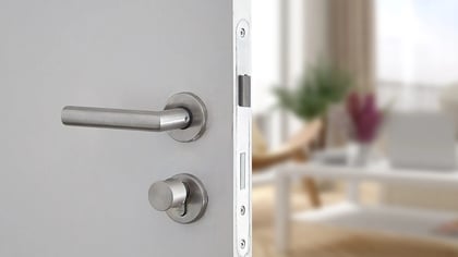 Manija y cerradura redonda de metal cepillado en una puerta blanca, ligeramente abierta a una sala de estar difuminada.