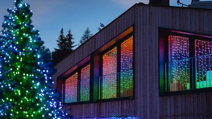 Bunte LED-Lichterketten schmücken ein modernes Holzhaus und einen Weihnachtsbaum in winterlicher Abendlandschaft.