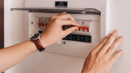 Una mano con reloj acciona los interruptores naranjas de un panel de disyuntores blanco, mostrando su uso.