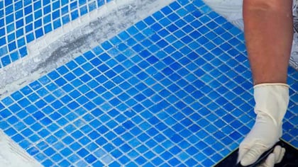 Persona con guantes instalando azulejos de mosaico azul brillante en una superficie, posiblemente una piscina o baño.