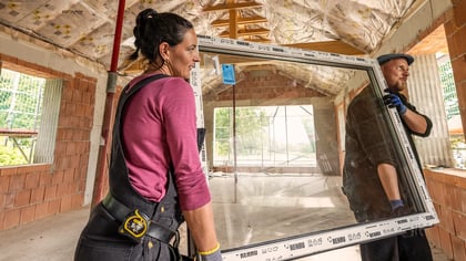 Dos personas transportan un gran marco de ventana REHAU blanco con cristal en un edificio en construcción.