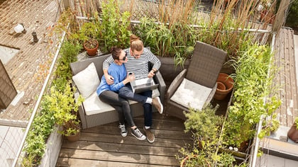 Zwei Personen entspannen auf einem modernen Outdoor-Sofa auf einer hölzernen Dachterrasse, umgeben von Pflanzen und nutzen einen silbernen Laptop.