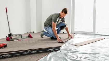 Hombre instalando paneles de suelo laminado efecto madera clara en una habitación, con herramientas cerca.