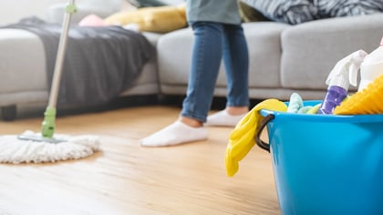 Blauer Putzeimer gefüllt mit gelben Handschuhen, Sprühflasche, Bürste und Tüchern, auf Holzboden. Im Hintergrund wischt eine Person den Boden.
