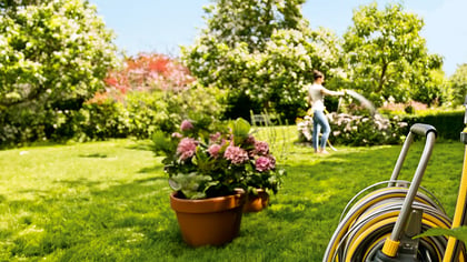 Gelbe Gartenschlauchaufroller, der an einer Holzwand befestigt ist, auf dem Rasen mit Blumen und einer Frau im Hintergrund.