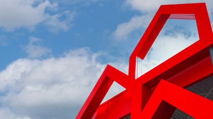 Estructura de acero pintado de rojo contra cielo azul con nubes, formando un punto arquitectónico destacado.