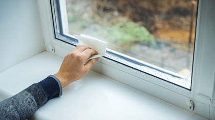 Una mano usa una toalla de papel blanca para limpiar la condensación de un marco de ventana blanco.