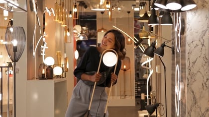 Mujer sonriente sostiene lámpara de pie moderna con marco de madera clara y luz esférica blanca en una tienda.