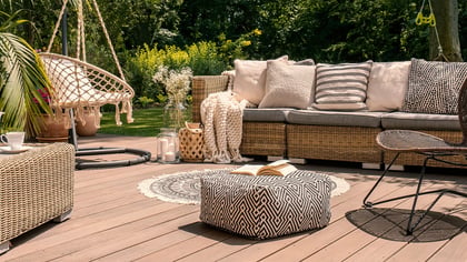 Ein einladender Außenbereich mit Korbmöbeln, darunter Sofa, Couchtisch und Sitzgelegenheiten, auf Holzterrasse inmitten üppigen Grüns.
