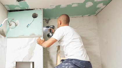 Ein Handwerker befestigt mit einem Akkuschrauber eine weiße Marmorfliese an einer Wand in einem Badezimmer. Die Fliese hat eine graue Maserung.
