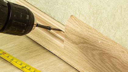 Bohrmaschine schraubt Schraube in Sockelleiste, daneben ein gelbes Maßband auf hellem Boden. Werkzeug und Baumaterial im Detail.