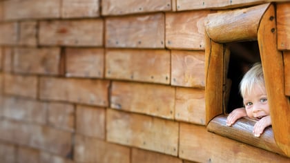 Ein kleines Kind späht durch ein Holzfenster und lächelt, das Gesicht umrahmt von blonden Haaren und die Hände auf der Fensterbank.