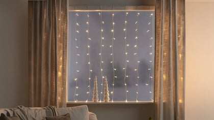 Gemütliches Wohnzimmer mit Vorhängen, Lichterketten und dekorativen Weihnachtsbäumen auf der Fensterbank für festliche Stimmung.