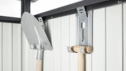 Eine Nahaufnahme von Gartengeräten wie einer Schaufel und einem Hammer, die ordentlich an einer weißen Wand aufgehängt sind.