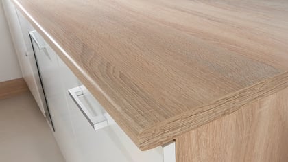Nahaufnahme einer Küchenarbeitsplatte aus hellem Holz mit weißen Schrankfronten und Griffen. Die Holzmaserung ist gut sichtbar.