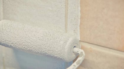 Un rodillo de pintura aplicando pintura blanca sobre azulejos marrones claros.