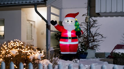 Beleuchteter aufblasbarer Weihnachtsmann im roten Anzug, einen kleinen Weihnachtsbaum haltend, im Schnee stehend.