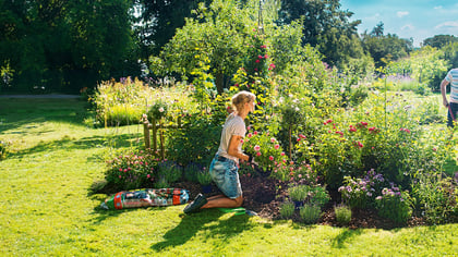 Ein Garten mit einer Frau, die kniet und gärtnert, während ein Mann mit einer Schubkarre voller Blumen im Hintergrund steht.