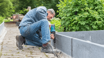 Mann installiert graue Beeteinfassungen/Hochbeetelemente entlang eines Weges im Garten.