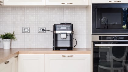 Vista de la cocina con una cafetera automática negra sobre una encimera clara, integrada en una cocina moderna con armarios blancos.