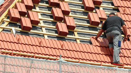 Rote Dachziegel werden von einem Arbeiter auf einem Holzdachstuhl verlegt, mit neuen Ziegelstapeln.