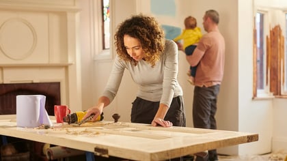 Mujer quitando pintura de una puerta de madera con una herramienta eléctrica, en una habitación en renovación con su familia.