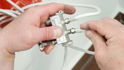 Manos conectan cables coaxiales blancos a un divisor de cable plateado.