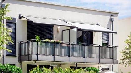 Zwei ausfahrbare graue Markisen über einem modernen Balkon mit dunklem Metallgeländer und Milchglas.