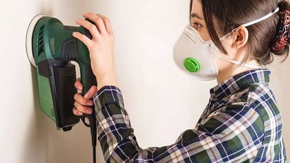Persona con camisa de cuadros y mascarilla usa una lijadora orbital verde y negra para alisar una pared.